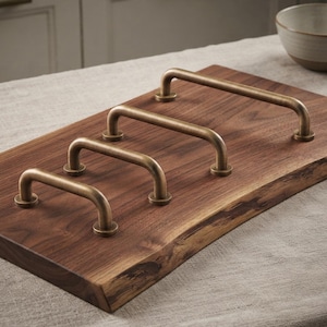 May include: Four U-shaped cabinet pulls with a brushed antique brass finish are mounted in a diagonal row on a rectangular piece of dark brown walnut wood with a natural live edge, showcasing a smooth grain and rich, deep wood tones.