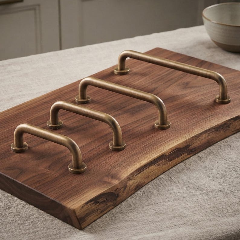 May include: Four U-shaped cabinet pulls with a brushed antique brass finish are mounted in a diagonal row on a rectangular piece of dark brown walnut wood with a natural live edge, showcasing a smooth grain and rich, deep wood tones.