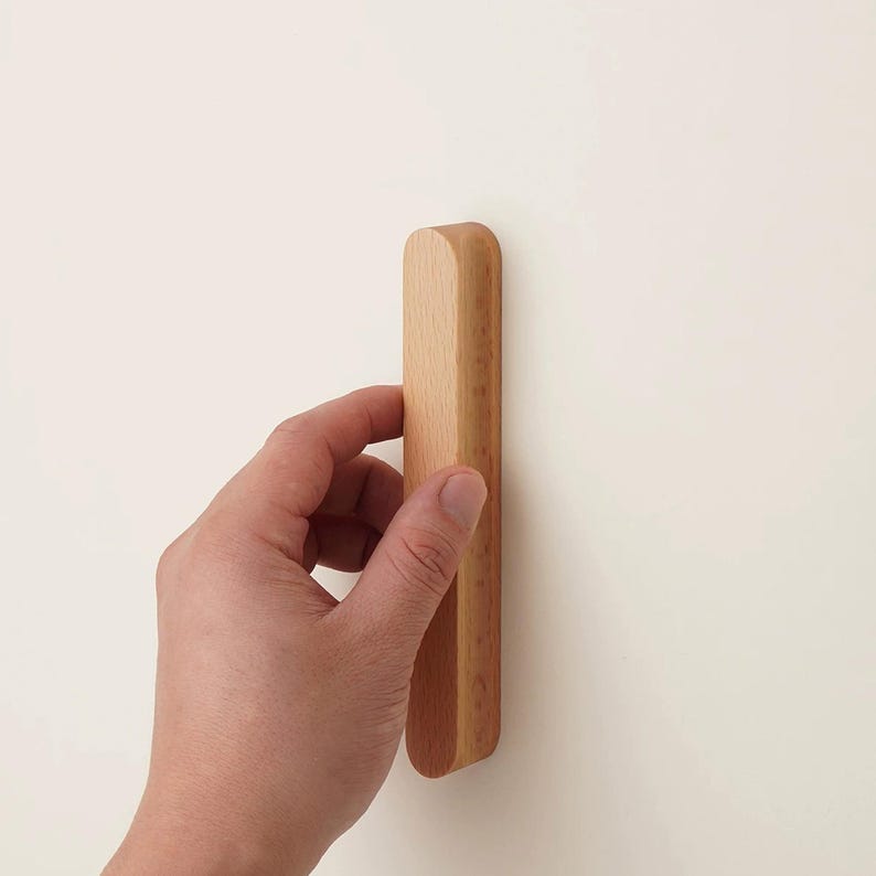 May include: A light-coloured, natural wood cabinet handle with a smooth, elongated rectangular shape and rounded edges. The handle features a visible wood grain pattern and a matte finish, designed for attachment to a flat surface.