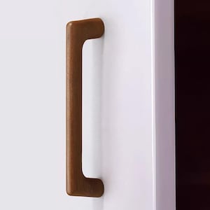 May include: A vertical, rectangular wooden cabinet handle with rounded edges and a smooth, medium-brown finish. The handle is mounted onto a flat, white surface, featuring a simple, solid construction with a consistent grain texture throughout.