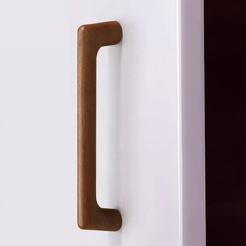 May include: A vertical, rectangular wooden cabinet handle with rounded edges and a smooth, medium-brown finish. The handle is mounted onto a flat, white surface, featuring a simple, solid construction with a consistent grain texture throughout.