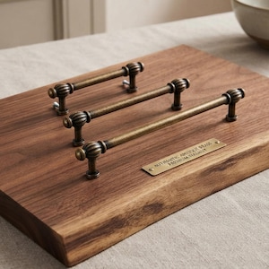 Pode incluir: Três puxadores de armário de latão antigo com extremidades caneladas montados numa peça retangular de madeira de nogueira castanha escura. Uma pequena placa de latão está fixada na madeira com o texto gravado: AUTHENTIC ANTIQUE BRASS PREMIUM WALNUT.
