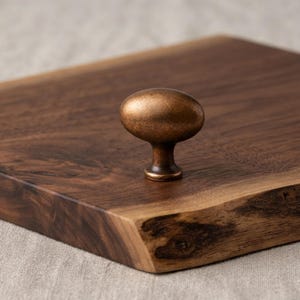 May include: An oval-shaped cabinet knob with a brushed bronze finish mounted on a dark brown wooden board featuring a natural live edge. The knob has a smooth, rounded top and a tapered base, showcasing a metallic texture and warm brown tones.