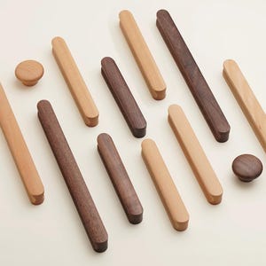 May include: A collection of wooden cabinet hardware including various lengths of rectangular drawer pulls and two circular knobs. The hardware features a mix of light-toned wood and dark-toned wood finishes with smooth, rounded edges and a simple design.