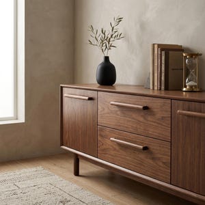 May include: A dark wood sideboard featuring two side cabinet doors and two central drawers with horizontal wooden handles. On top sits a matte black vase with green branches, a small stack of books, and a gold-toned hourglass timer.