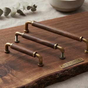 May include: Three cabinet pulls of varying lengths featuring cylindrical dark brown wood grips and brushed brass metal ends. A small rectangular brass plaque is attached to the wood surface with the text Solid Walnut & Aged Brass Collection Premium Hardware.