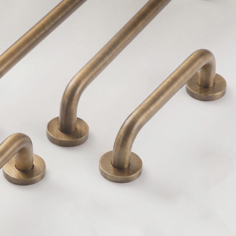 May include: A set of three brushed antique brass cabinet pulls, each featuring a smooth, rounded U-shaped handle attached to two circular mounting bases. The metal hardware has a warm, metallic finish with subtle linear texture throughout the surface.