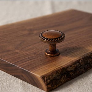 May include: A round, antique bronze-toned metal cabinet knob featuring a decorative twisted rope border around the edge. The knob is mounted on a rectangular piece of dark brown wood with a natural, rough bark edge on one side.