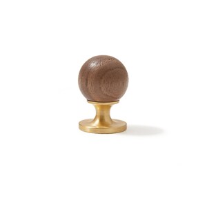 May include: A spherical brown wooden knob attached to a circular brushed brass base. The wood features a visible natural grain pattern, while the metal base has a smooth, matte finish. This hardware component stands upright on its flat bottom.