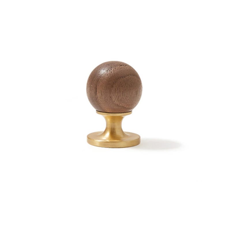 May include: A spherical brown wooden knob attached to a circular brushed brass base. The wood features a visible natural grain pattern, while the metal base has a smooth, matte finish. This hardware component stands upright on its flat bottom.