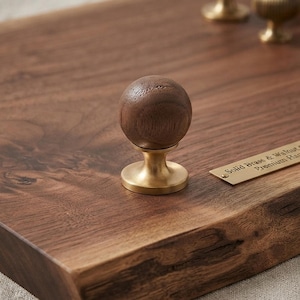 Puede incluir: Un pomo esférico de madera marrón oscuro montado sobre una base circular de latón cepillado. El pomo está fijado a una gruesa losa de madera de nogal oscuro con un borde natural. Una pequeña placa rectangular de latón tiene grabado Solid Brass & Walnut.