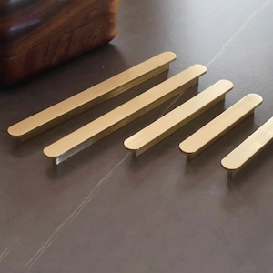 Könnte beinhalten: Fünf goldfarbene Schrankgriffe aus Metall, die in einer diagonalen Reihe angeordnet sind. Jeder Griff hat einen langen, schlanken, rechteckigen Körper mit abgerundeten Enden und einer glatten, gebürsteten Metalloberfläche. Die Griffe variieren in der Länge.
