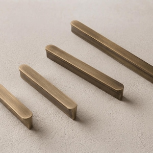 Peut inclure: Quatre poignées de placard linéaires avec une finition en laiton antique brossé, disposées en diagonale de la plus courte à la plus longue. Chaque poignée présente une surface supérieure arrondie et lisse avec un profil rectangulaire.