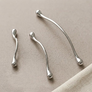 Peut inclure: Trois poignées de placard en métal argenté avec une finition lisse et réfléchissante. Chaque poignée présente une forme incurvée et allongée avec des extrémités arrondies et bulbeuses, variant en longueur d'une pièce courte à un arc long.