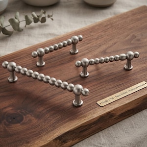 May include: Three satin nickel cabinet pulls with a beaded design, each featuring a series of spheres along the handle. A small rectangular metal plaque is attached to the wood, displaying the text: Model 2700 - Satin Nickel - Premium Walnut.