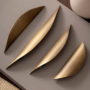 May include: Four curved cabinet pulls with a brushed brass finish. Each handle features a tapered, elongated crescent shape with a smooth, metallic texture. The pulls are arranged diagonally, showcasing their varying lengths and pointed ends.