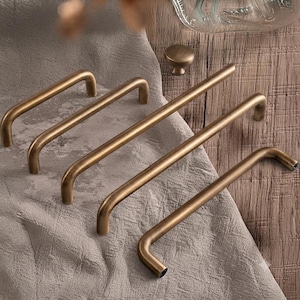 May include: A collection of four brushed brass cabinet pulls with a smooth, rounded U-shaped design and one matching circular cabinet knob. The hardware features a warm, metallic finish with a consistent cylindrical profile throughout each piece.