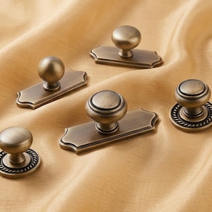 May include: A collection of five antique brass cabinet knobs and pulls. Three knobs feature rectangular backplates with scalloped edges, while two knobs are circular with a rope-textured base. All pieces have a brushed metallic finish.