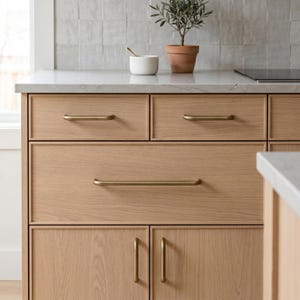 May include: A light oak kitchen cabinet unit featuring two top drawers, one large middle drawer, and two lower cabinet doors. Each drawer and door is fitted with a slim, horizontal or vertical brushed brass handle. The unit has a white countertop.
