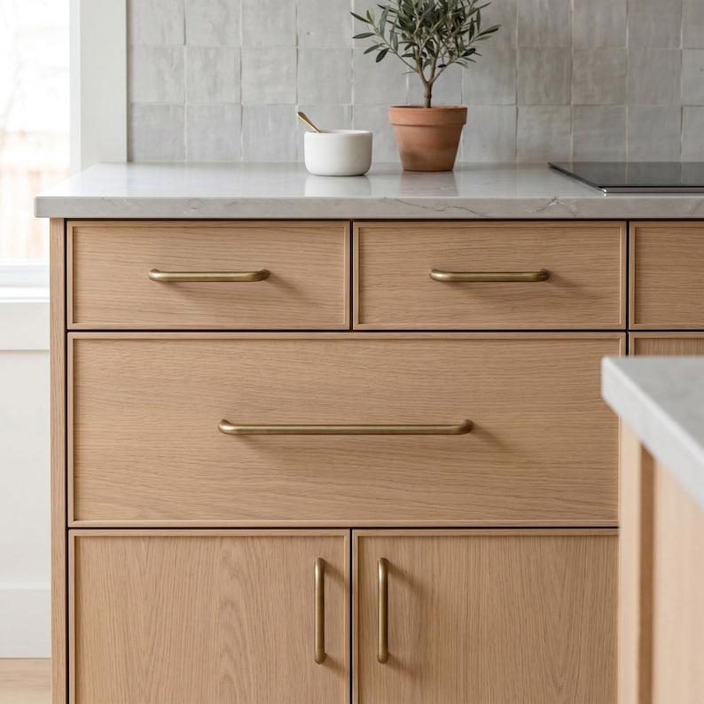 May include: A light oak kitchen cabinet unit featuring two top drawers, one large middle drawer, and two lower cabinet doors. Each drawer and door is fitted with a slim, horizontal or vertical brushed brass handle. The unit has a white countertop.
