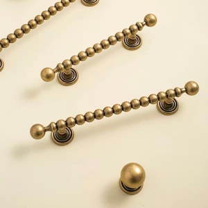 May include: A collection of antique brass-toned cabinet hardware including three elongated drawer pulls with a beaded design and one matching spherical knob. Each piece features a circular base and a textured metallic finish.