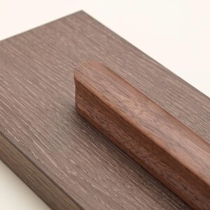 May include: A rectangular piece of dark brown wood with a visible grain pattern featuring a smooth, rounded, elongated wooden handle attached to the surface. The handle is a slightly darker shade of brown than the base, showcasing natural wood texture.
