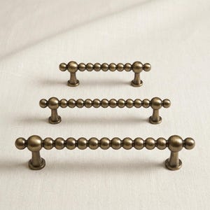 May include: Three antique brass cabinet pulls arranged in a vertical row, each featuring a horizontal bar composed of a series of connected spheres and supported by two small, flared pedestals at the ends.