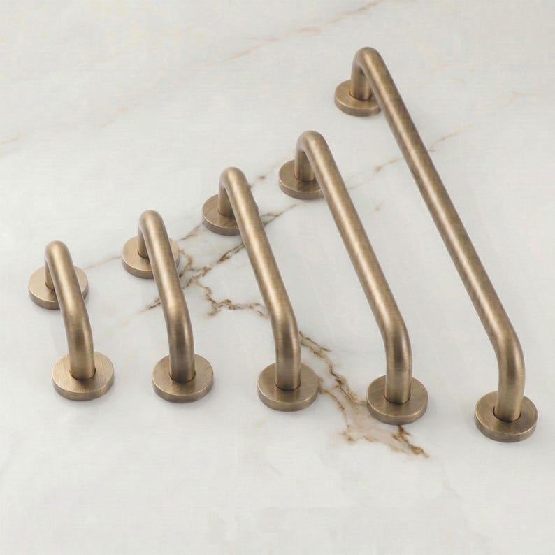 May include: A set of five arch-shaped cabinet pulls in a brushed brass finish, arranged in a diagonal line from smallest to largest. Each handle features a smooth, rounded bar design with two circular mounting bases at the ends for attachment.