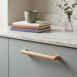 May include: A light wood cabinet handle with a smooth, cylindrical shape attached to a matte grey cabinet door. The handle features a natural grain pattern and rounded ends, providing a simple, functional grip for opening the cabinet door.