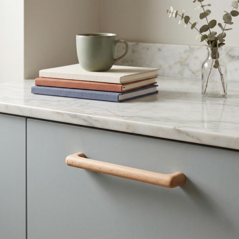 May include: A light wood cabinet handle with a smooth, cylindrical shape attached to a matte grey cabinet door. The handle features a natural grain pattern and rounded ends, providing a simple, functional grip for opening the cabinet door.