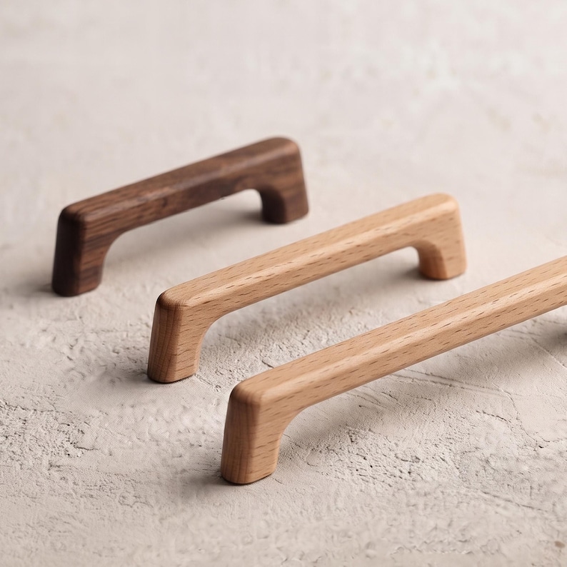 May include: Three wooden cabinet pulls arranged in a diagonal line. The back pull is dark brown with a visible wood grain, while the two front pulls are a lighter, natural tan wood colour. Each handle features a smooth, curved arch design.