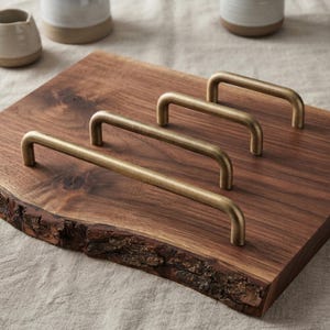 May include: Three brushed brass cabinet pulls of varying lengths arranged diagonally on a dark brown wooden board with a natural live edge. The metal handles feature a simple, rounded U-shaped design with a smooth, matte metallic finish.