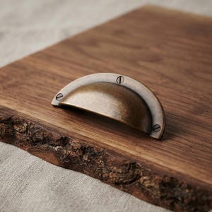 May include: A semi-circular cup drawer pull with an antique brass finish and a smooth, curved surface. The hardware features three visible screw holes, each containing a flat-head screw, and is mounted onto a dark brown wooden board.