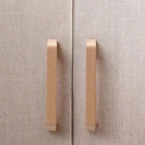 May include: Two vertical light-colored wooden cabinet handles mounted on a pair of doors covered in a textured beige fabric. The handles have a smooth, rectangular profile with slightly angled ends, contrasting against the woven material.