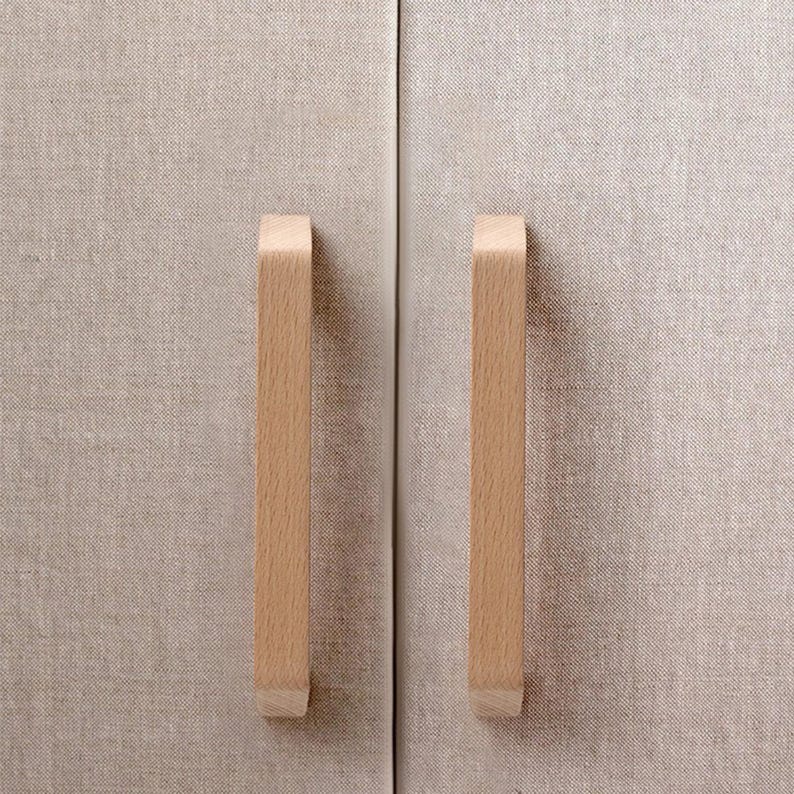 May include: Two vertical light-colored wooden cabinet handles mounted on a pair of doors covered in a textured beige fabric. The handles have a smooth, rectangular profile with slightly angled ends, contrasting against the woven material.