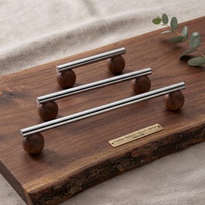 May include: Three cabinet pulls featuring polished silver-tone cylindrical bars mounted on spherical dark brown walnut wood bases. A small rectangular brass plaque is attached to the wood, engraved with the text: Premium Walnut & Chrome Collection - Handcrafted.