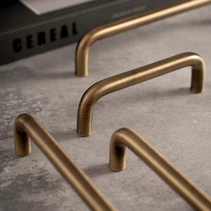 May include: Several U-shaped cabinet pulls with a brushed antique brass finish. Each handle features a smooth, rounded profile and two mounting points. The metal hardware displays a subtle, linear texture across the surface of the curved bars.