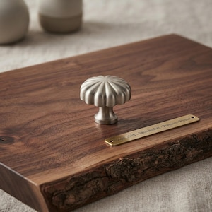 May include: A satin nickel cabinet knob with a fluted, circular design mounted on a dark brown walnut wood board. A small brass-colored metal plate is attached to the wood, featuring the engraved text: Model 2309 - Satin Nickel - Premium Walnut.