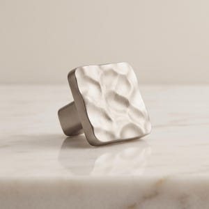 May include: A square cabinet knob with a brushed silver finish and a textured, hammered surface. The knob features a rounded square face with irregular indentations across the front, creating a reflective, metallic appearance for furniture hardware.