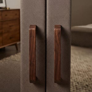 May include: Two vertical, rectangular panels upholstered in a textured, taupe-colored fabric. Each panel features a long, smooth, dark brown wooden handle mounted vertically in the center, providing a contrast to the fabric surface.