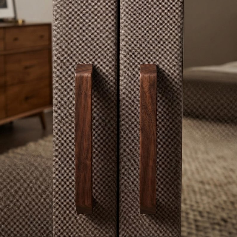 May include: Two vertical, rectangular panels upholstered in a textured, taupe-colored fabric. Each panel features a long, smooth, dark brown wooden handle mounted vertically in the center, providing a contrast to the fabric surface.