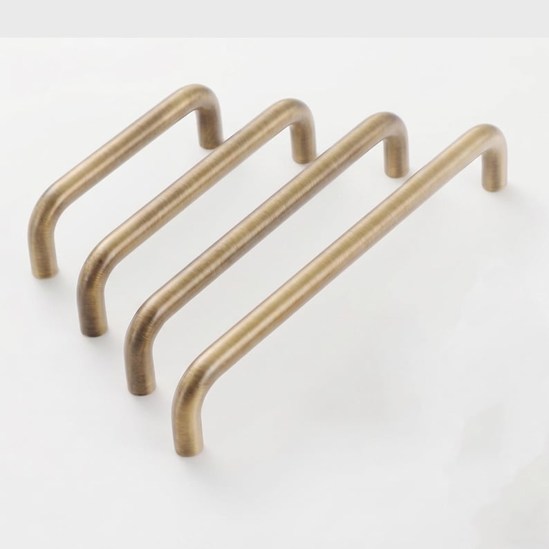 May include: Four metal cabinet pulls with a brushed antique brass finish. Each handle features a simple, rounded U-shaped design with two mounting points at the ends. The pulls are arranged in a row, showing varying lengths from shortest to longest.