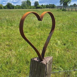 Op de afbeelding: Een hartvormige metalen sculptuur, gemonteerd op een verweerde houten paal. De sculptuur heeft een roestbruine kleur, tegen een achtergrond van een groen veld, blauwe lucht en pluizige witte wolken. Een prikkeldraadomheining staat op de voorgrond.