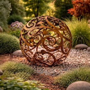 Op de afbeelding: Een decoratief, bolvormig metalen tuinornament met een ingewikkeld, opengewerkt ontwerp van wervelende patronen. De roestbruine bol staat te midden van een bed van kleine stenen, groene planten en grote rotsen, wat een natuurlijke, buitenesthetiek creëert.