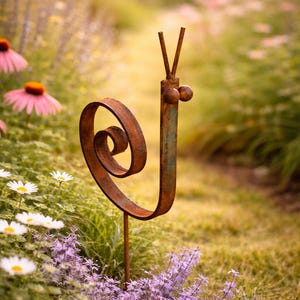 Könnte beinhalten: Eine rostige Schnecken-Gartenfigur aus Metall mit einem eingerollten Gehäuse und Fühlern steht in einem Garten. Die Skulptur ist braun mit einem Hauch von Grün, umgeben von rosa Sonnenhüten, weißen Gänseblümchen und lila Blumen.