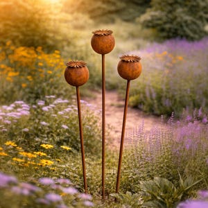 Op de afbeelding: Drie roestige metalen tuinsculpturen in de vorm van klaprozen. Ze staan in een tuin vol paarse en gele bloemen. Elke sculptuur heeft een ronde zaadkop met een getextureerde bovenkant en een lange steel.