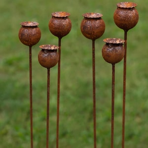 Puede incluir: Seis estacas de jardín de metal oxidado, cada una con un diseño de cabeza de semilla esférica. Las cabezas de las semillas tienen una apariencia texturizada y desgastada, y están montadas sobre tallos verticales y delgados. Las estacas están dispuestas sobre un fondo verde borroso.