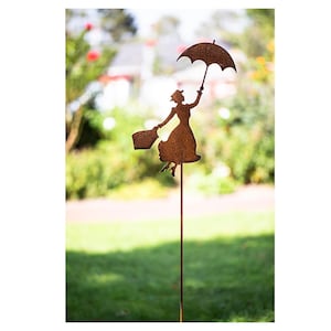 Può includere: Un paletto da giardino in metallo marrone arrugginito con la sagoma di una donna che tiene un ombrello e una borsa, come se stesse fluttuando. L'ornamento da giardino è posto in un giardino erboso con uno sfondo sfocato di alberi e una casa.