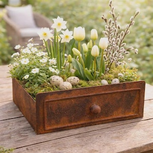 Peut inclure: Un tiroir en métal rouillé de forme rectangulaire rempli de mousse verte, de jonquilles blanches, de tulipes blanches, de branches de saule marsault et de petites fleurs blanches. Plusieurs œufs tachetés sont nichés dans la mousse.