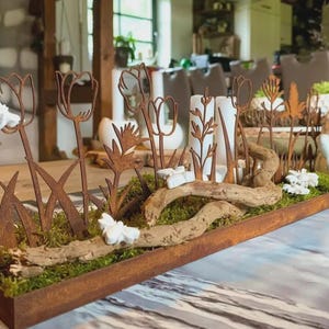 Op de afbeelding: Een rustiek, rechthoekig tafelstuk met metalen tulp- en plantuitsparingen, witte kaarsen en een keramische kikker. De opstelling omvat mos, drijfhout en witte decoratieve elementen, alles in een verweerde houten bak.